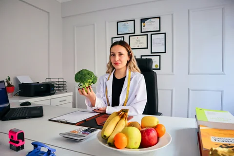 Ernährungsberaterin im weißen Kittel mit Maßband in ihrer Praxis, Brokkoli in der Hand und Obstschale auf dem Schreibtisch