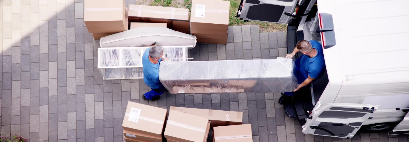 Zwei Umzugshelfer in blauer Arbeitskleidung tragen ein in Folie verpacktes Sofa zu einem weißen Transporter, daneben gestapelte Umzugskartons